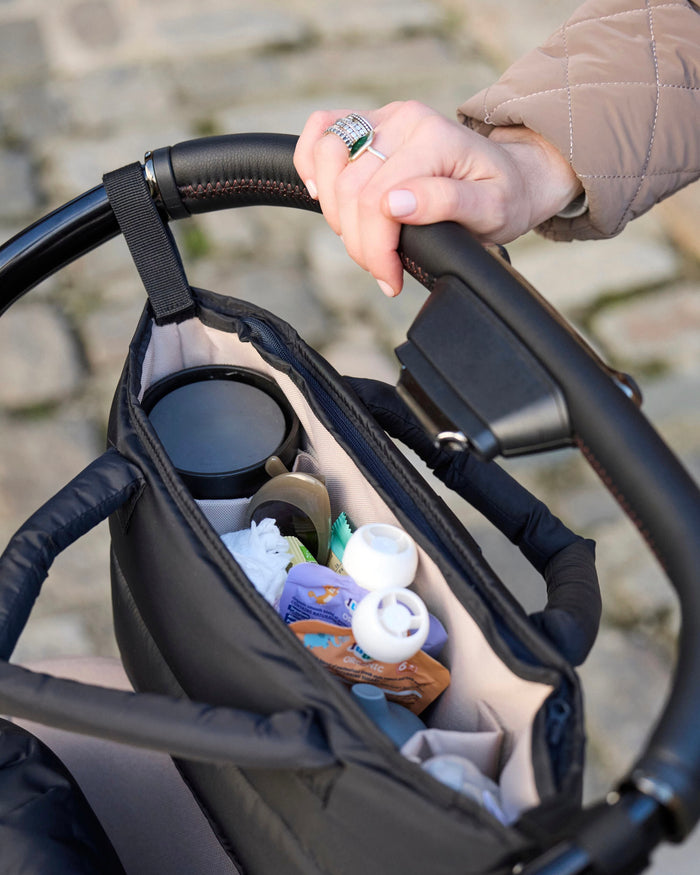 Hand holding a stroller handle with a storage bag containing items.