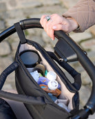 Hand holding a stroller handle with a storage bag containing items.