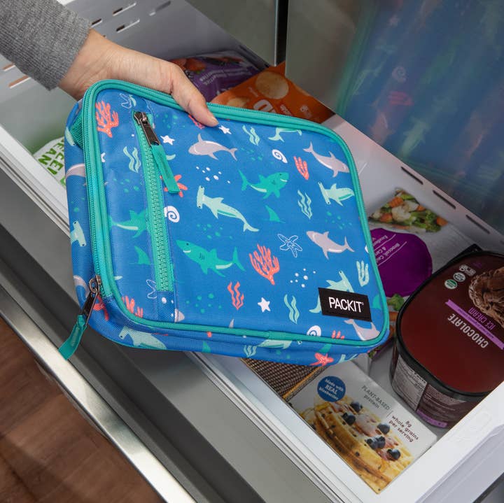 Blue insulated lunch bag with shark design held in front of an open refrigerator.