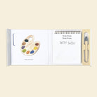 A children's coloring book or activity pad shown open on a light beige background. The left page features a watercolor-style paint palette with multiple colors and the text 'Name your color!'.