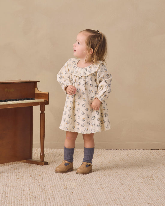 A toddler girl stands in a studio setting wearing a cream-colored long-sleeve dress with a delicate blue floral pattern. She is styled with dark gray socks and tan shoes, standing next to a wooden piano against a warm beige background.