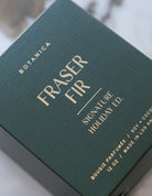 Green packaging of Fraser Fir candle with gold text on a blurred background