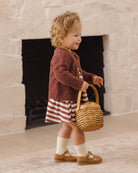 A toddler stands in front of a stone fireplace holding a small woven basket. The child wears a rust-brown knit cardigan over a red-and-white striped dress, white socks, and tan Mary Jane shoes.