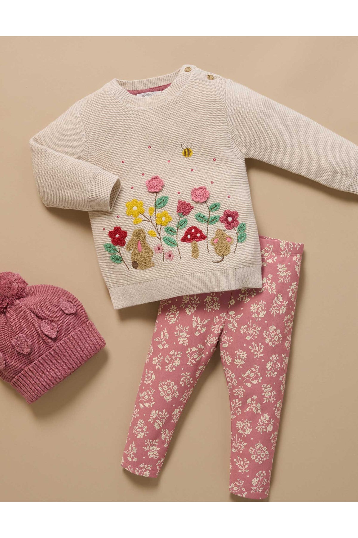 A baby girl's two-piece knit outfit set featuring a cream-colored long-sleeve sweater with colorful embroidered flowers, mushrooms, and a bunny, paired with pink leggings with a white floral pattern. A matching pink knit pom-pom hat is also shown on a solid beige background.