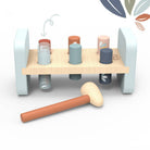 A wooden toy peg-banging game with a light natural wood base, pale blue side supports, colorful cylindrical pegs, and a matching wooden mallet on a white background.