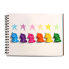 Six colorful unicorn-shaped crayons in blue, purple, pink, orange, yellow, and green are lined up on a white spiral notebook page below matching colored star crayons.