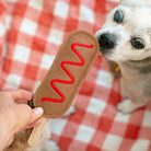 A small dog licks a brown felt hot dog toy with a red wavy line, held by a hand against a red and white checkered blanket.