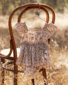 A baby girl's short-sleeve romper and matching shorts set in a beige and brown floral print, hanging on a wooden hanger from the back of a rustic wooden chair in a dry grass field.