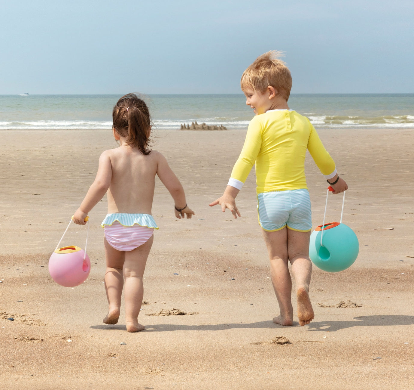 Two children walking on a beach with sand toys.