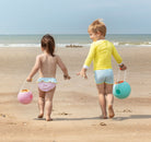 Two children walking on a beach with sand toys.
