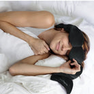 Person lying in bed with a black sleep mask on, wearing a white shirt.