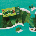 The Crissis Green Jumbo Puzzle box placed on top of a partially assembled green leaf collage puzzle, with a few loose puzzle pieces scattered around on a turquoise textured background.