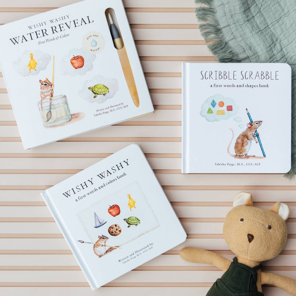 Two children's picture books are arranged on a light wood surface with a soft green fringed textile and a plush teddy bear. The books are titled 'Wishy Washy Water Reveal' and 'Scribble Scrabble'.