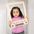 Child holding a sign saying 'my first day of school' framed by a wooden picture frame.