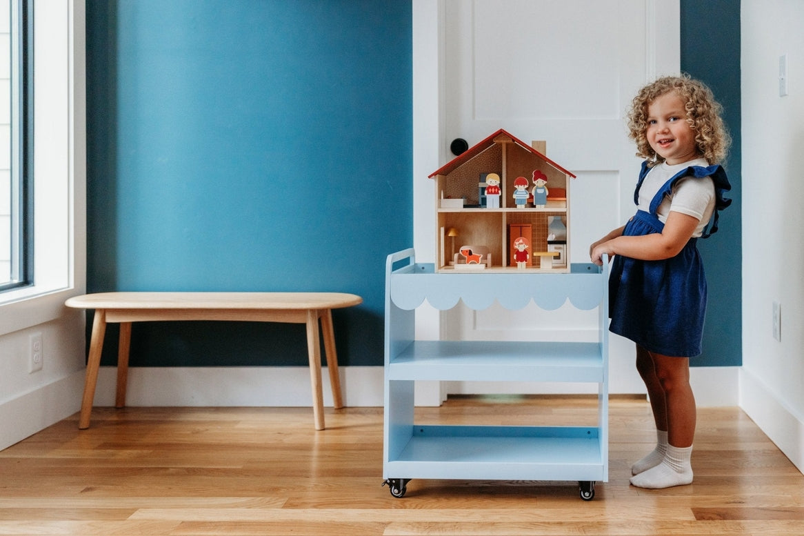 A young child with curly brown hair standing beside a blue rolling utility cart, reaching toward an open-faced wooden dollhouse by Mentari filled with miniature furniture.