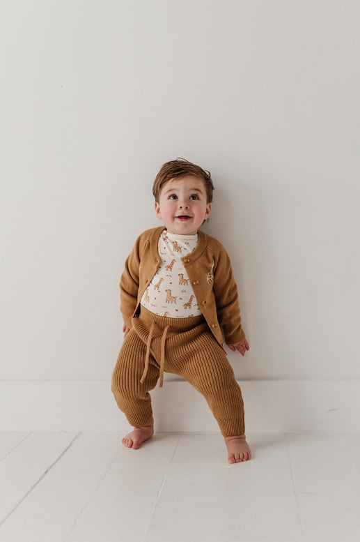 A baby wearing a matching brown knit cardigan and pants set over a white patterned bodysuit, standing barefoot against a plain white wall.