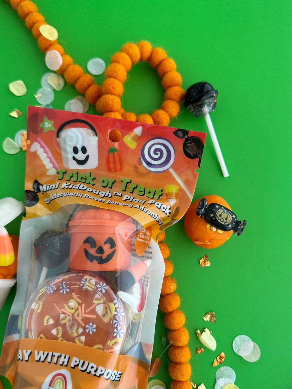 A packaged Halloween mini craft set on a bright green background, surrounded by candy and orange pom-pom garlands. The package contains a jack-o'-lantern shaped cup, a spider decoration, and a small orange pumpkin with a black witch hat.