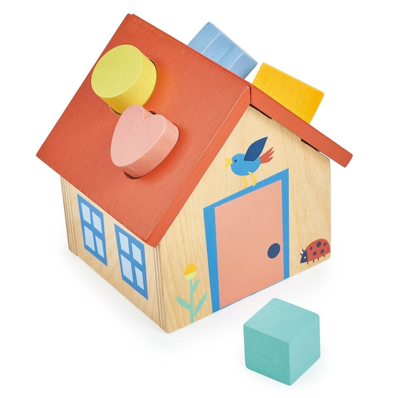 A charming wooden shape sorter toy designed as a little house with a red roof, featuring shape-sorting slots on the roof and front facade. A blue bird decoration sits on the roof, a flower pot stands near the front door, and a ladybug is illustrated near the double windows. Several chunky, brightly colored wooden pieces—including a yellow cylinder, a light blue cross, a mint green cube, a yellow triangle, and a pink heart—rest on the white surface in front of the house.