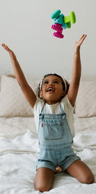 A young child sitting on a bed, smiling and reaching up toward the Knudll Sensory Activity Toy by Mobi Games floating above them.