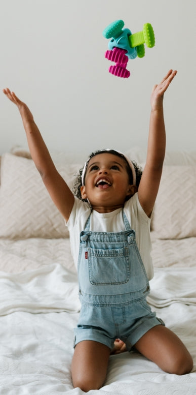 A young child sitting on a bed, smiling and reaching up toward the Knudll Sensory Activity Toy by Mobi Games floating above them.