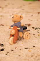 A small plush teddy bear wearing a knitted blue sweater sits on a wooden toy scooter with natural wood wheels and handlebars, placed on a beige carpet.
