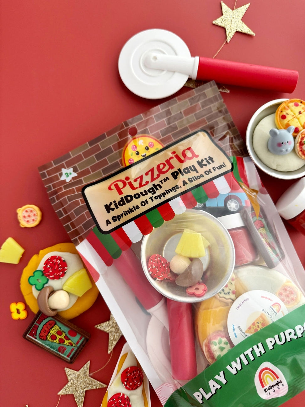 Pizzeria KidDough play kit in a clear plastic bag with a brick-patterned header, surrounded by small toy pizza and food-themed accessories on a red background.