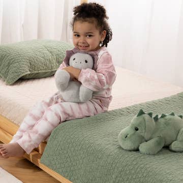 A young child wearing pink and white striped pajamas sits on a bed holding a gray plush toy, with a green quilted blanket and matching pillow in the background.