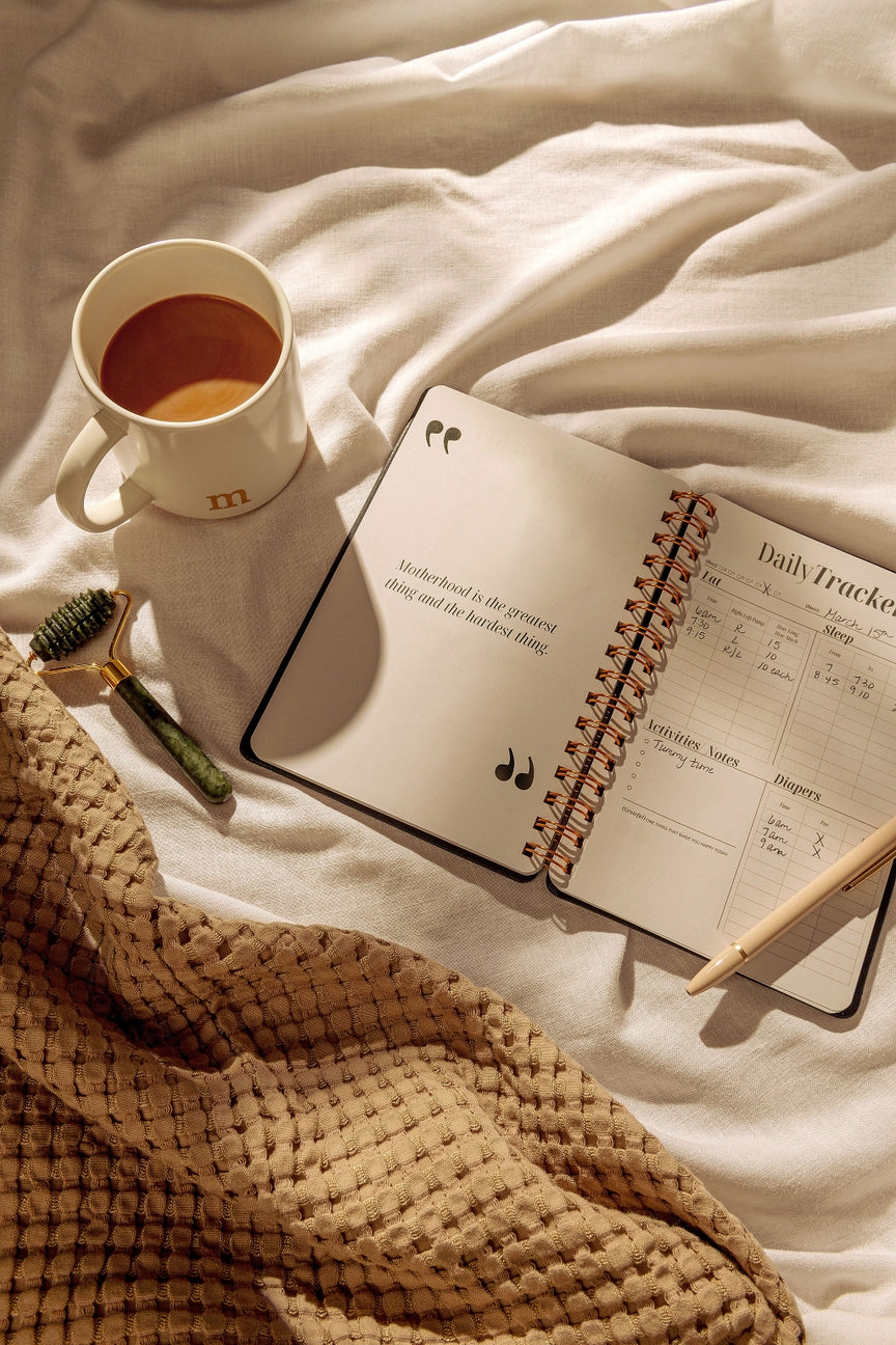 Open notebook with daily tracker on a bed next to a cup of coffee and a pen.