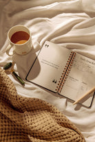 Open notebook with daily tracker on a bed next to a cup of coffee and a pen.
