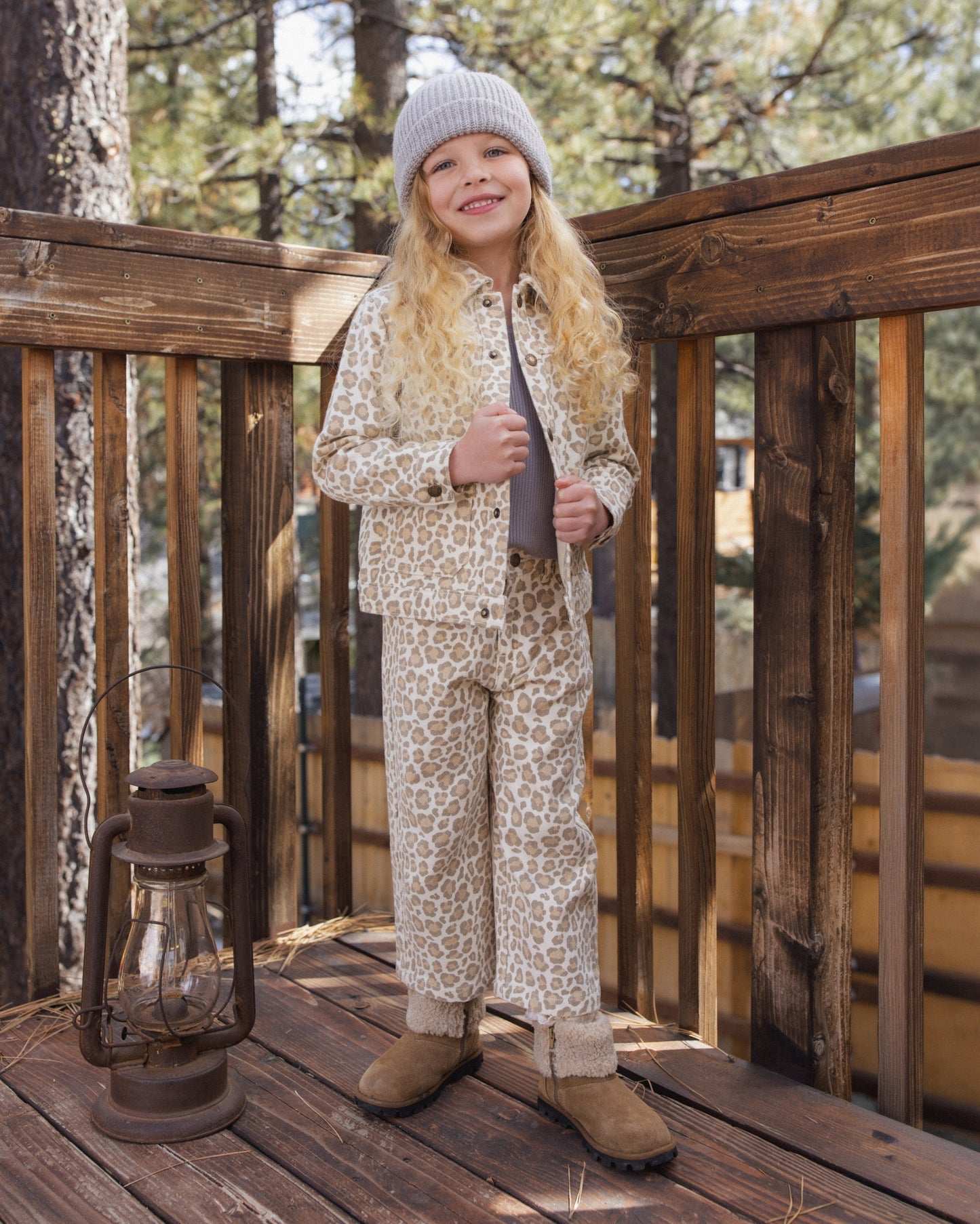 A young girl stands on a wooden deck wearing a matching beige and brown animal-print pajama set with a gray zip-up hoodie, a gray knit beanie, and brown boots. A rustic lantern and trees are visible in the background.