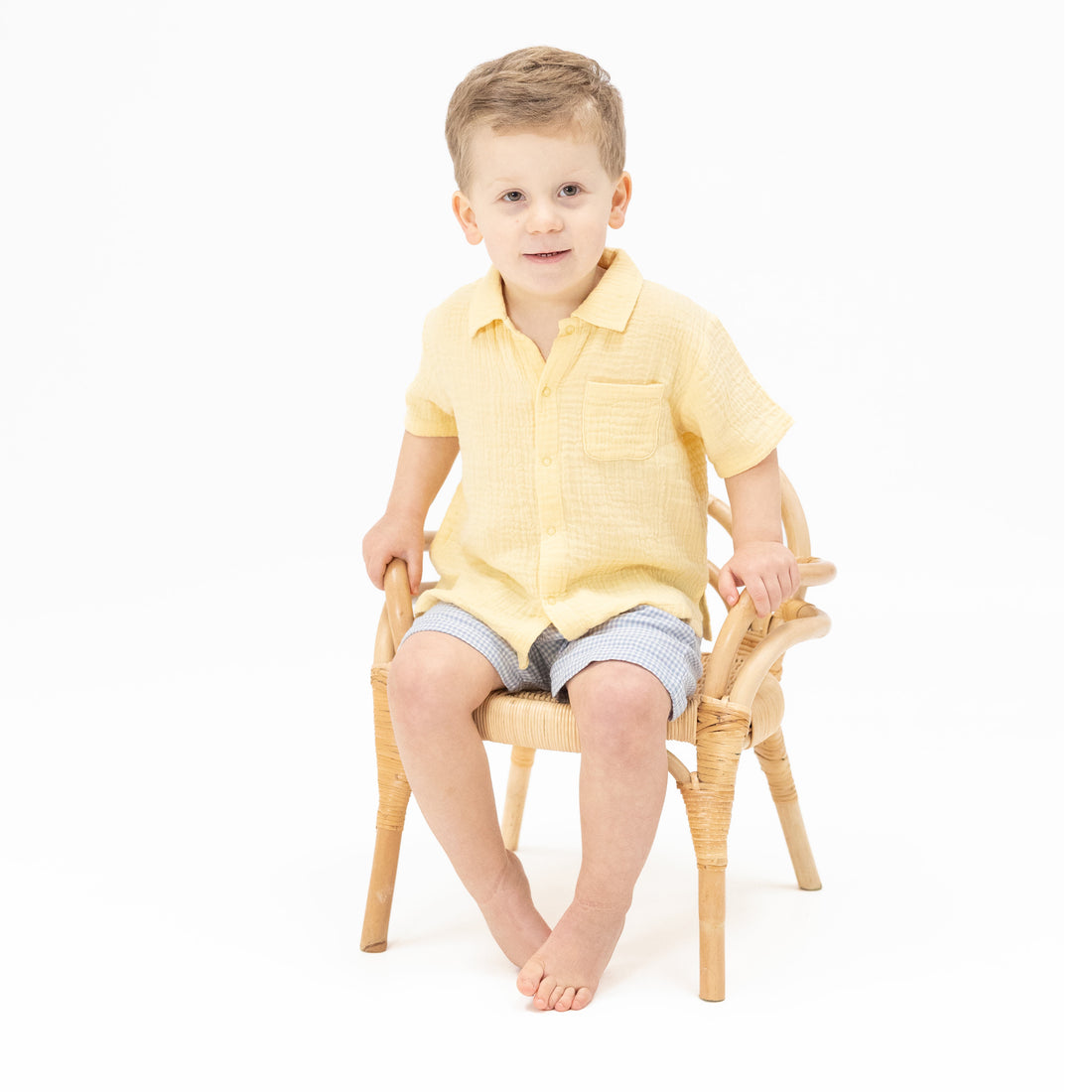 Young boy sitting on a small wooden chair wearing a yellow shirt and gray shorts.