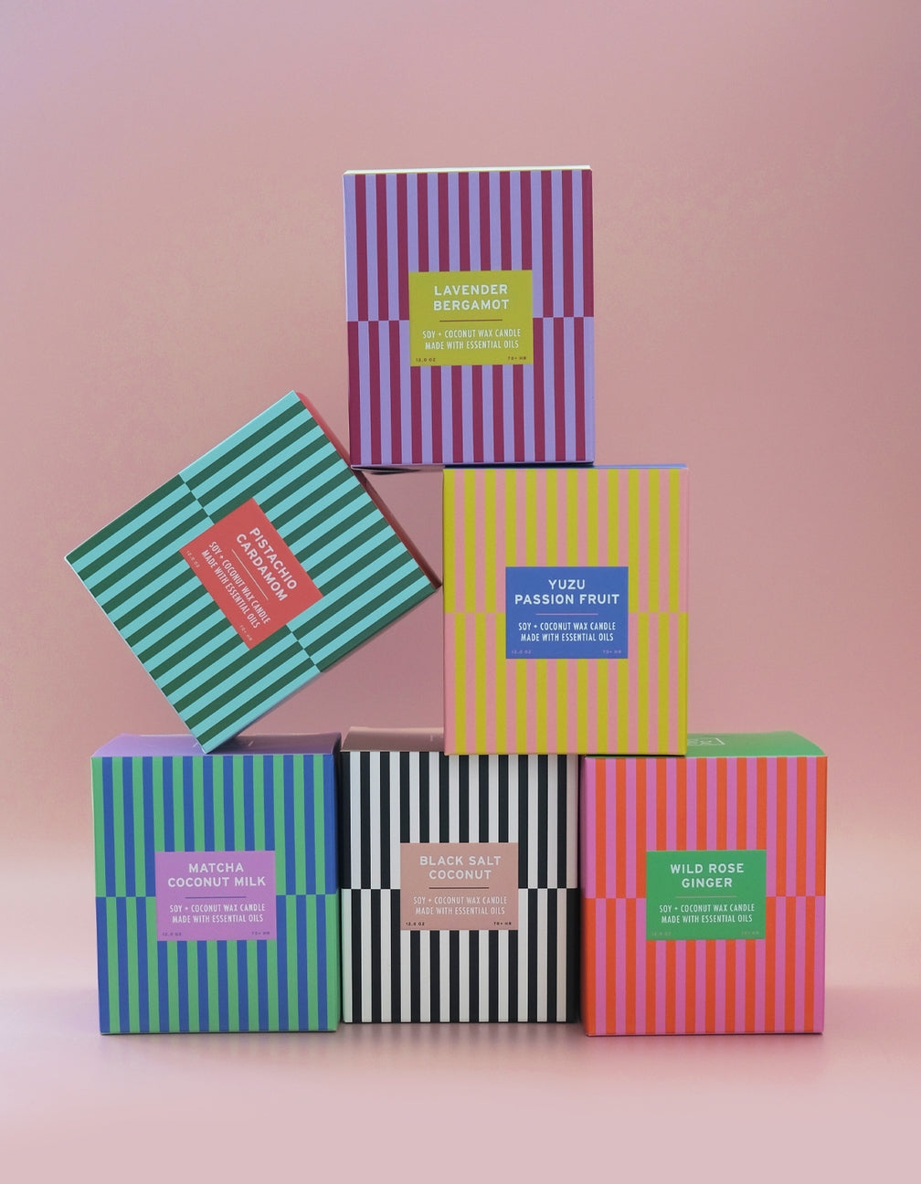 Six colorful striped boxes of scented candles arranged in a pyramid stack on a soft pink background. The boxes feature different color schemes and labels for scents including Lavender Bergamot, Yoga Passion Fruit, Matcha Coconut Milk, Black Salt Coconut, and Wild Rose Osmanthus.