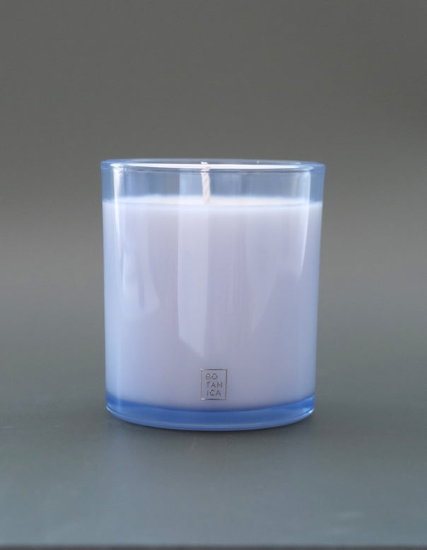 A white soy wax candle in a clear glass jar with a light blue tint, set against a solid gray background. The brand name 'SO TAN CO.