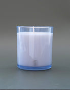 A white soy wax candle in a clear glass jar with a light blue tint, set against a solid gray background. The brand name 'SO TAN CO.