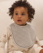 A close-up of a baby wearing a gray and white striped bib over a cream-colored patterned long-sleeve romper, set against a soft, warm off-white background.