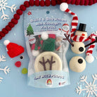 A packaged craft play set labeled 'Build a Snowman Mini Dough Play Dough' shown on a light blue background with festive snowflake decorations and red pom-pom garland. The clear bag contains snowman-themed dough pieces, and nearby are small red-and-white knit hats, a red-and-white striped scarf, and a small snowman figure made from dough.