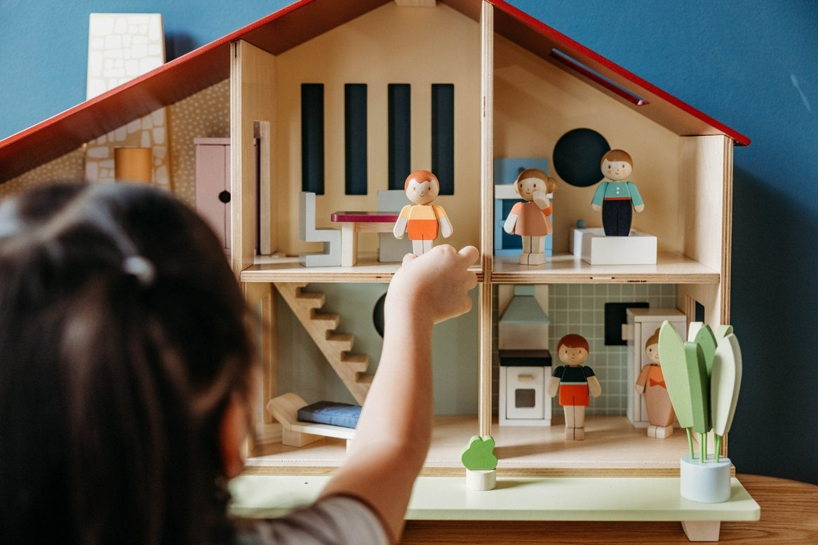 A child's hand reaches toward the open-front Wooden Cedar Chalet Modernist Dollhouse by Mentari, with miniature figures and furniture arranged across its six rooms and patio.
