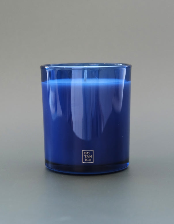 A cobalt blue glass candle jar with a white square logo at the bottom, photographed against a neutral gray background.