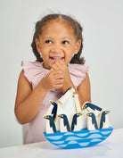 A young child with braided hair and a ruffled pink top smiling playfully while sitting next to the wooden balancing penguins and rocking iceberg toy.