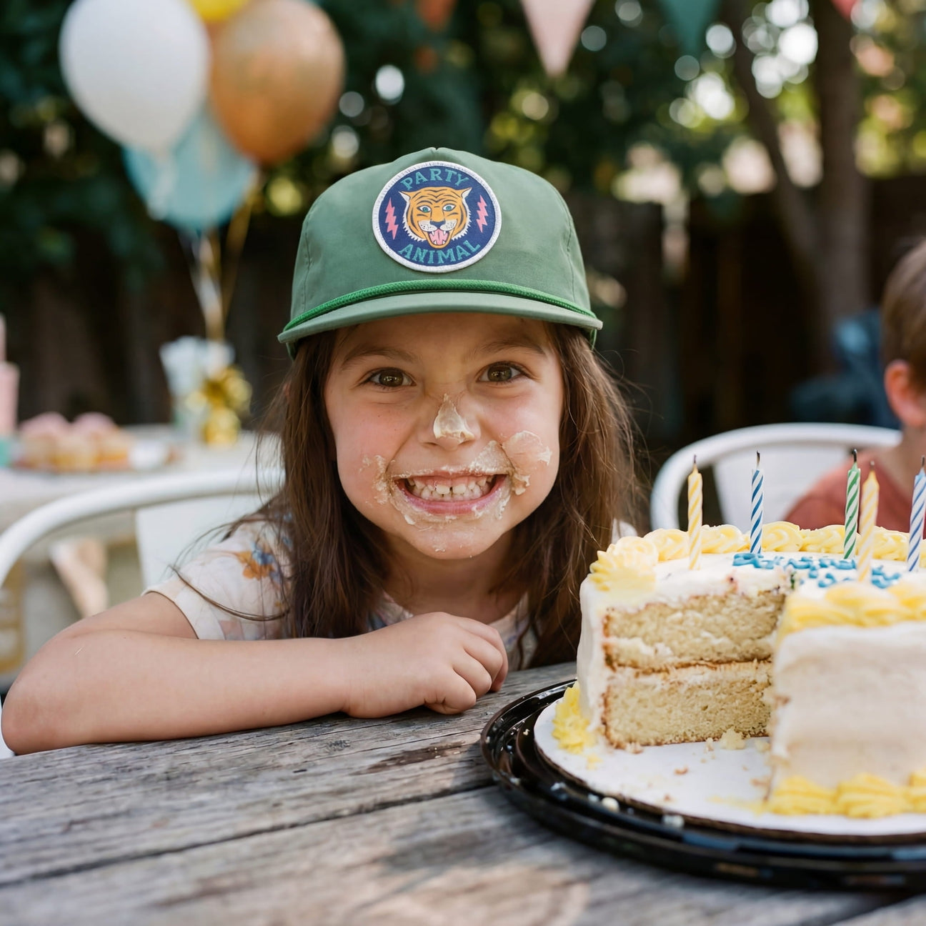 Child with cake batter on face, wearing a green cap, sitting at a table with a birthday cake.