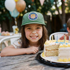 Child with cake batter on face, wearing a green cap, sitting at a table with a birthday cake.