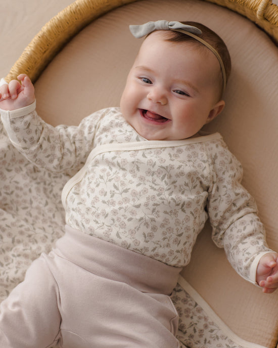 A smiling baby lying on their back in a woven bassinet, wearing a cream and beige floral long-sleeve wrap top with matching beige pants and a gray bow headband.