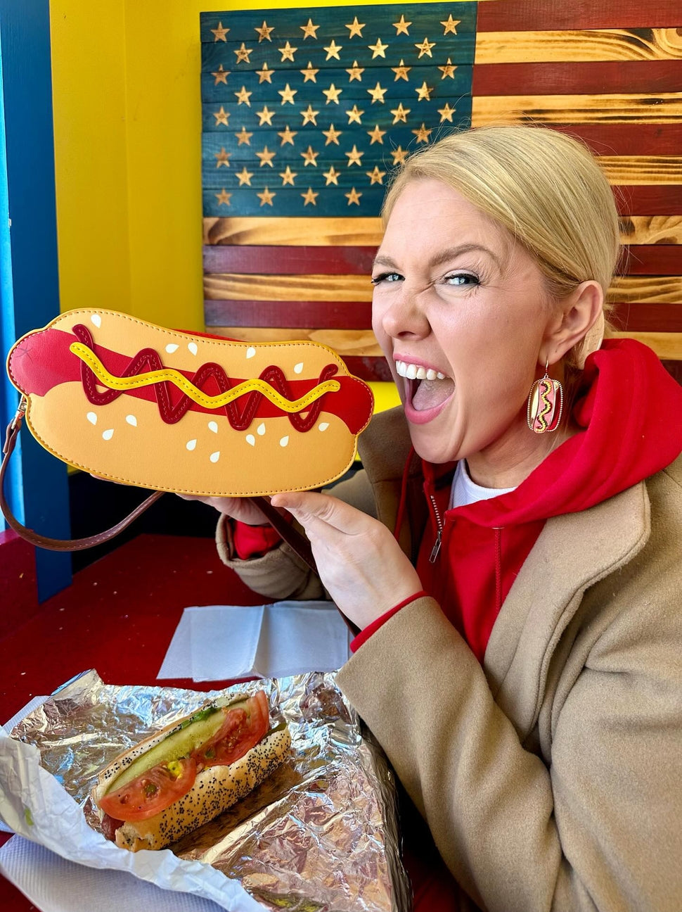 Person holding a hot dog-shaped bag with a sandwich on a table in front of an American flag.