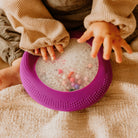 Child's hands interacting with a purple ring filled with colorful beads on a textured surface.