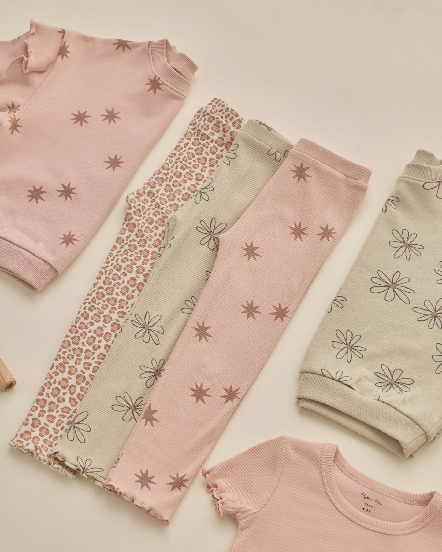 A flat-lay collection of children's pajamas in soft pastel tones, including a pink long-sleeve top with star prints, patterned leggings, and a beige top with daisy prints, arranged on a light cream background.