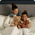 Woman and child sitting on a bed with a teddy bear, smiling.