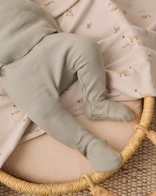 A close-up of a baby's legs wearing muted sage green waffle-knit booties, resting on a patterned cream muslin blanket inside a woven rattan bassinet.