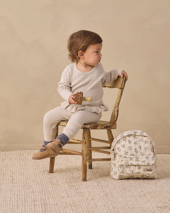 A toddler sits on a rustic wooden chair in a studio setting, wearing a cream ribbed long-sleeve top and matching cream leggings. A small cream backpack with a blue floral pattern rests beside the chair.