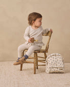 A toddler sits on a rustic wooden chair in a studio setting, wearing a cream ribbed long-sleeve top and matching cream leggings. A small cream backpack with a blue floral pattern rests beside the chair.