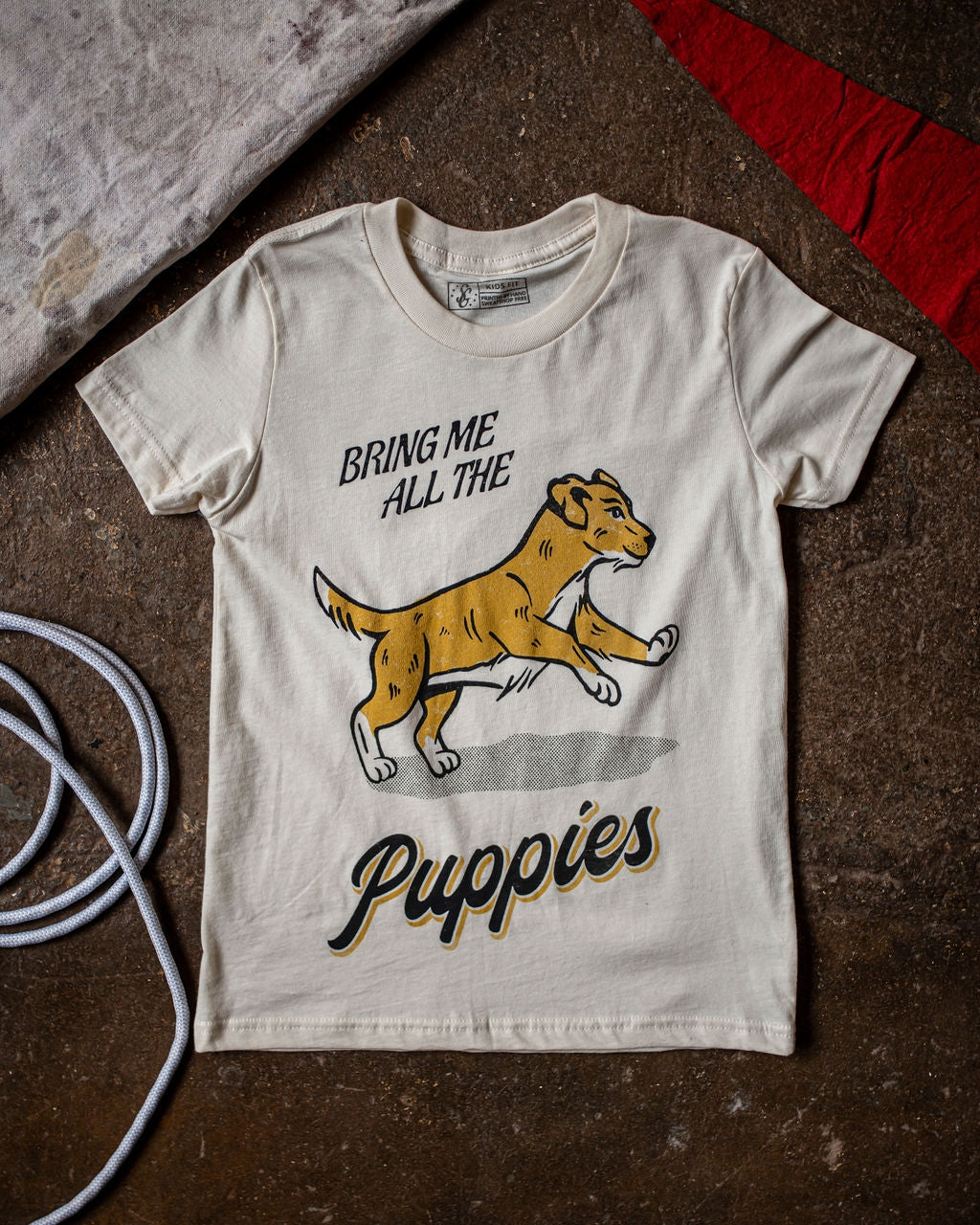 An off-white short-sleeve t-shirt,
featuring a graphic of a running puppy with the text 'BRING ME ALL THE Puppies' in brown.